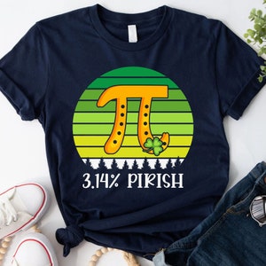 Puede incluir: Camiseta azul marino con un dise&ntilde;o circular verde y amarillo con el s&iacute;mbolo Pi en naranja, una herradura y un tr&eacute;bol. El texto "3.14% PIRISH" est&aacute; impreso debajo. Una divertida camiseta tem&aacute;tica del D&iacute;a de San Patricio.