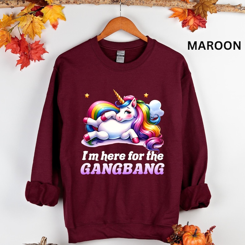 K&ouml;nnte beinhalten: Ein maroonfarbener Pullover mit einem wei&szlig;en Einhorn mit einer Regenbogenm&auml;hne und einem Regenbogen-Schwanz. Das Einhorn liegt auf dem Boden und ist von Sternen umgeben. Der Text "I'm here for the GANGBANG" ist in Lila unter dem Einhorn aufgedruckt.
