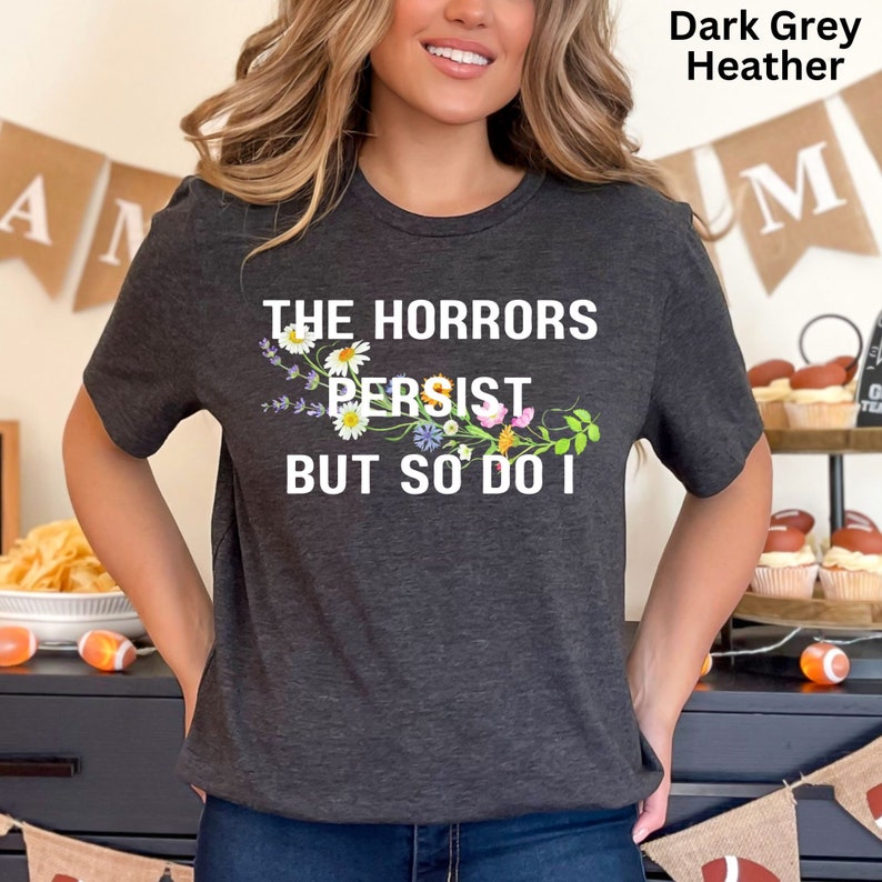 Puede incluir: Camiseta de color gris oscuro jaspeado con un dise&ntilde;o floral y el texto "The Horrors Persist But So Do I".