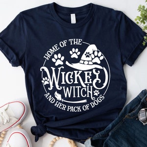Może przedstawiać: Granatowa koszulka z kr&oacute;tkim rękawem z białym nadrukiem graficznym o treści "HOME OF THE WICKED WITCH AND HER PACK OF DOGS", zawierająca ilustracje kapelusza czarownicy, odcisk&oacute;w łap i miotły ułożone w okrągły wz&oacute;r.