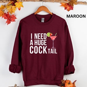 May include: A maroon sweatshirt with the text "I need a huge cocktail" in white letters and a cocktail glass with a lime and strawberry on the rim.