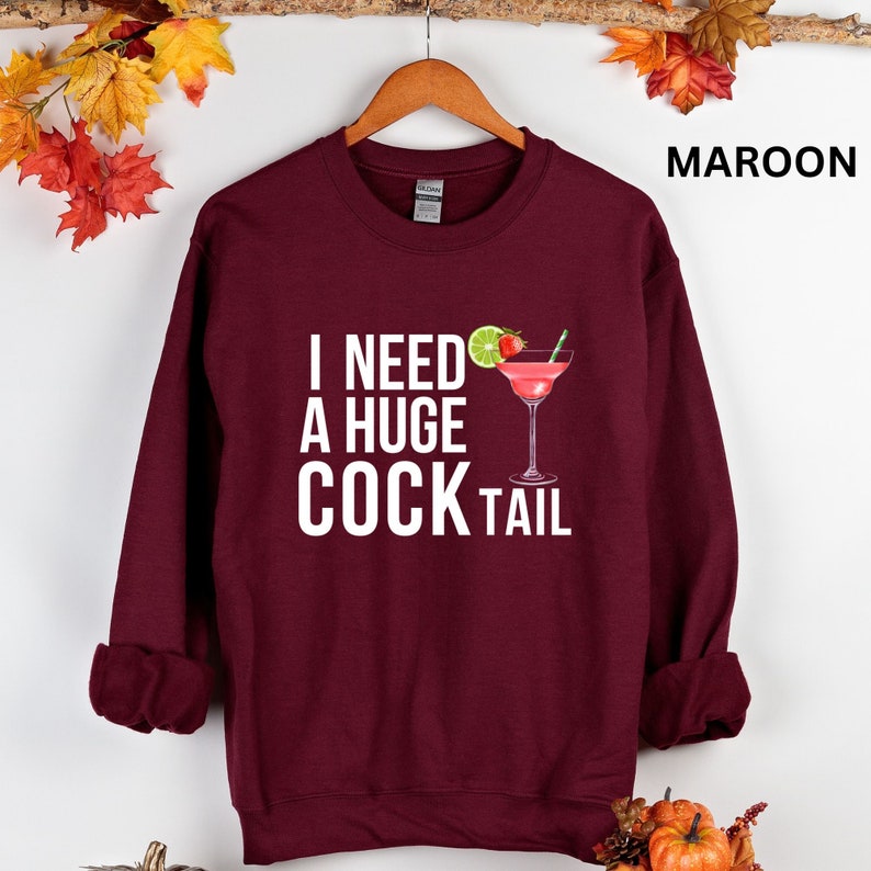 May include: A maroon sweatshirt with the text "I need a huge cocktail" in white letters and a cocktail glass with a lime and strawberry on the rim.
