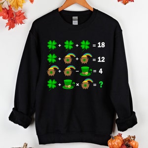 Puede incluir: Sudadera negra con un problema de matemáticas de temática del Día de San Patricio. El problema presenta tréboles, ollas de oro y sombreros de duende. La ecuación es:  + + = 18, + + = 12, + - = 4, + * = ?