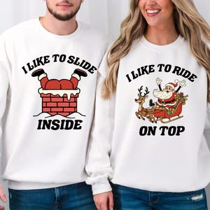 Könnte beinhalten: Weiße Sweatshirts mit weihnachtlichen Motiven. Eines zeigt den Weihnachtsmann, der einen Schornstein hinunterrutscht, mit dem Text "I LIKE TO SLIDE INSIDE". Das andere zeigt den Weihnachtsmann in einem Schlitten mit dem Text "I LIKE TO RIDE ON TOP".