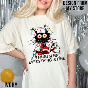 May include: A white t-shirt featuring a black cat wearing a nurse's hat and a Santa hat. The cat is tangled in Christmas lights and holding a stethoscope. The text on the shirt reads "It's fine. I'm fine. Everything is fine."
