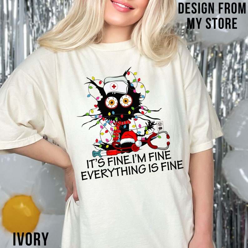 May include: A white t-shirt featuring a black cat wearing a nurse's hat and a Santa hat. The cat is tangled in Christmas lights and holding a stethoscope. The text on the shirt reads "It's fine. I'm fine. Everything is fine."