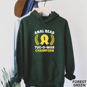 May include: A forest green hooded sweatshirt with the text "ANAL BEAD TUG-O-WAR CHAMPION" in white and yellow. The design includes a yellow ribbon and laurel wreath. The words "FOREST GREEN" are at the bottom right.