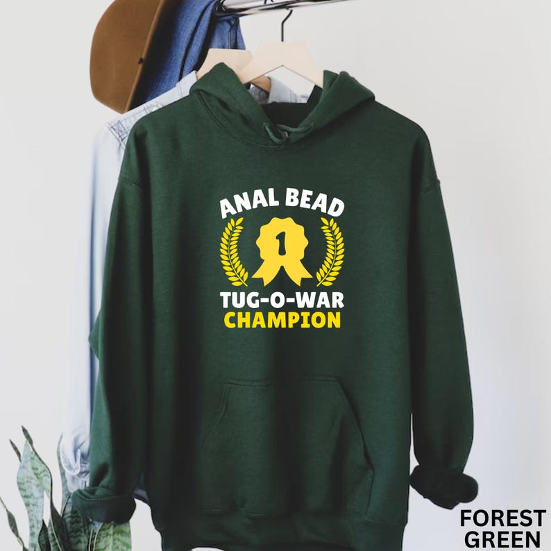 May include: A forest green hooded sweatshirt with the text "ANAL BEAD TUG-O-WAR CHAMPION" in white and yellow. The design includes a yellow ribbon and laurel wreath. The words "FOREST GREEN" are at the bottom right.