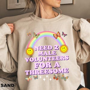 May include: A beige sweatshirt with a rainbow, two smiley faces, butterflies, and the text "Need 2 Male Volunteers For A Threesome".