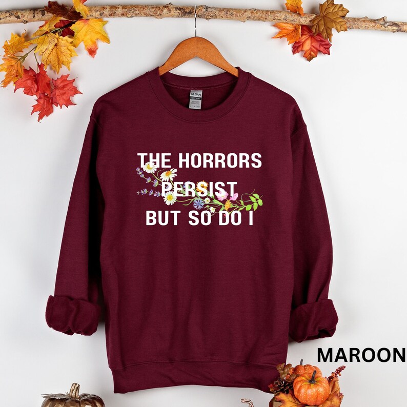 Puede incluir: Sudadera color granate con el texto "The Horrors Persist But So Do I" impreso en blanco con un dise&ntilde;o floral.