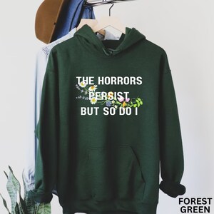 Puede incluir: Sudadera con capucha verde bosque con el texto "THE HORRORS PERSIST BUT SO DO I" impreso en blanco. El texto est&aacute; rodeado por un dise&ntilde;o floral.