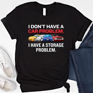 Puede incluir: Camiseta negra con el texto "I DON'T HAVE A CAR PROBLEM. I HAVE A STORAGE PROBLEM." La camiseta presenta ilustraciones de cuatro coches de diferentes colores: amarillo, blanco, azul y rojo.