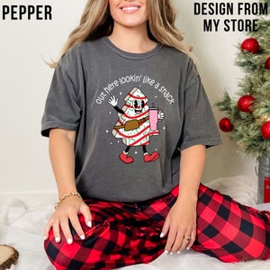 May include: A grey t-shirt featuring a cartoon Christmas tree wearing a candy cane pattern and holding a pink drink. The text on the shirt reads "Out here lookin' like a snack".
