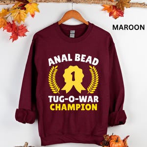 May include: Maroon sweatshirt with the text "ANAL BEAD TUG-O-WAR CHAMPION" in white and yellow. The design includes a yellow ribbon with the number 1 and a laurel wreath. The word "MAROON" is printed in white on the upper right.