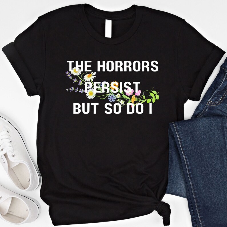 Puede incluir: Camiseta negra con texto blanco que dice "The Horrors Persist But So Do I". El texto est&aacute; rodeado por un dise&ntilde;o floral.