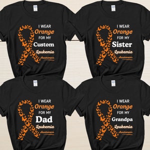 May include: Four black t-shirts with an orange ribbon and butterfly design. The text on the shirts reads "I wear orange for my [name] Leukemia Awareness". The names on the shirts are "Custom", "Sister", "Dad", and "Grandpa".