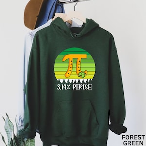 Puede incluir: Sudadera con capucha verde oscuro con un dise&ntilde;o gr&aacute;fico que presenta un s&iacute;mbolo Pi naranja dentro de un c&iacute;rculo a rayas verdes y amarillas, y el texto "3.14% PIRISH". El dise&ntilde;o incluye un tr&eacute;bol y peque&ntilde;os &aacute;rboles blancos.