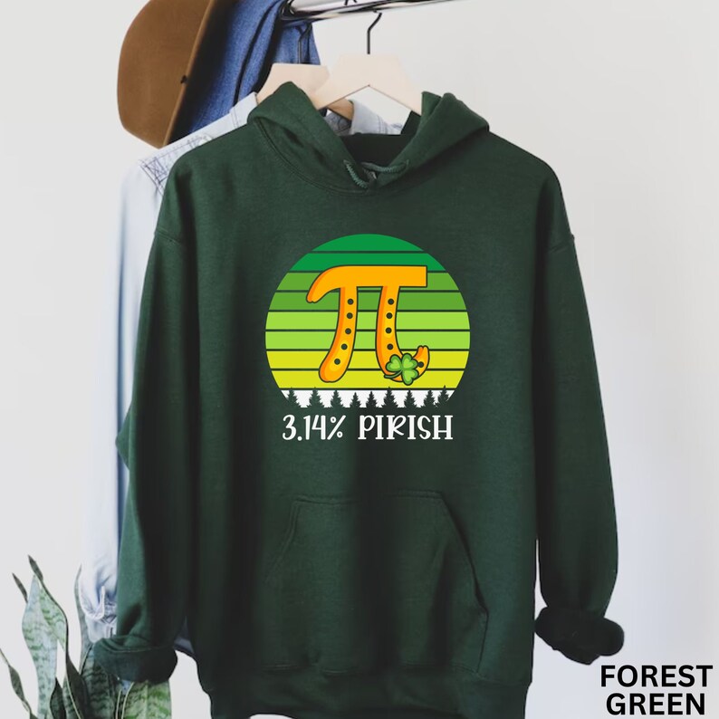 Puede incluir: Sudadera con capucha verde oscuro con un dise&ntilde;o gr&aacute;fico que presenta un s&iacute;mbolo Pi naranja dentro de un c&iacute;rculo a rayas verdes y amarillas, y el texto "3.14% PIRISH". El dise&ntilde;o incluye un tr&eacute;bol y peque&ntilde;os &aacute;rboles blancos.