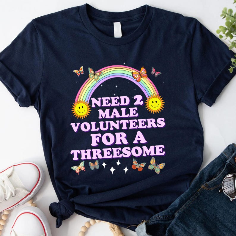 May include: A navy blue t-shirt with a rainbow, two suns, and butterflies. The text on the shirt reads "Need 2 Male Volunteers For A Threesome".