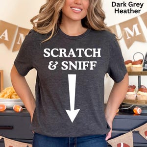 May include: A dark grey heather t-shirt with the text "SCRATCH & SNIFF" and a white arrow pointing down.