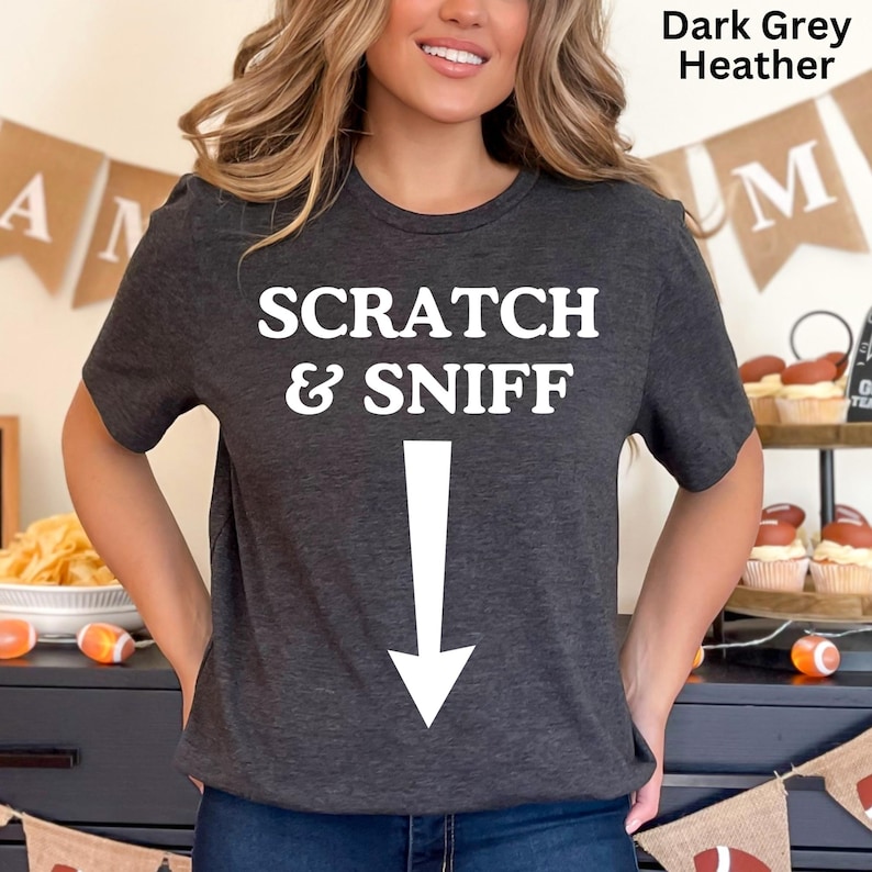 May include: A dark grey heather t-shirt with the text "SCRATCH & SNIFF" and a white arrow pointing down.