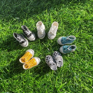 May include: A collection of eight miniature sneakers in various colours and patterns, including black and white chequered, yellow, blue, and white, arranged in a circle on a green grassy surface.