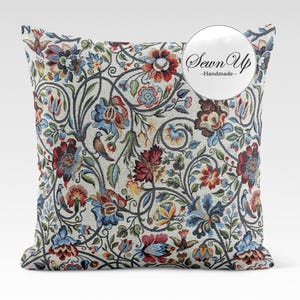 Può includere: Cuscino quadrato con un design arazzo floreale. Il cuscino presenta uno sfondo color crema con un motivo ripetuto di fiori rossi, blu e gialli, foglie verdi e viti grigio scuro. Il testo "Sewn Up - Handmade" è in un cerchio bianco.