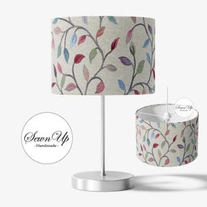 May include: A table lamp with a cylindrical lampshade. The lampshade has a beige background with a pattern of colorful leaves in red, blue, and purple. The lamp has a silver base and stem. The text "Sewn Up - Handmade" is in a circle.
