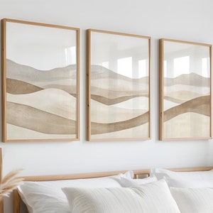 Könnte beinhalten: Ein weißes Bett mit einem weißen Bettbezug und einer beigefarbenen Strickdecke. Über dem Bett hängen drei gerahmte abstrakte Landschaftsdrucke mit braunen und beigefarbenen Aquarell-artigen Designs. Das Bett ist mit weißen Kissen und einem beigefarbenen Kissen mit einem dezenten Streifenmuster gestylt.