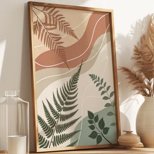 Op de afbeelding: Een ingelijste botanische kunstprint met illustraties van varens en bladeren in groen, bruin en beige tinten. Het kunstwerk wordt getoond op een houten kast, naast een vaas met gedroogd pampasgras, een glazen fles en een kaars.