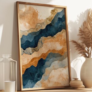 Può includere: Una stampa artistica astratta incorniciata con una cornice in legno. L'opera presenta forme a strati in stile acquerello nei toni del blu, dell'oro e del beige. La stampa è esposta su un mobile in legno, accanto a un vaso con piante secche e oggetti decorativi.