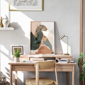 May include: A framed art print featuring a leopard illustration in shades of brown, green, and peach. The artwork is displayed on a wooden desk with a small potted plant, a desk lamp, and a wooden chair. A framed abstract art piece hangs above.