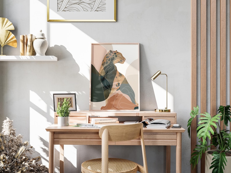 May include: A framed art print featuring a leopard illustration in shades of brown, green, and peach. The artwork is displayed on a wooden desk with a small potted plant, a desk lamp, and a wooden chair. A framed abstract art piece hangs above.