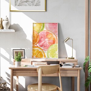 May include: A framed watercolor print of citrus fruit slices in shades of orange, pink, and green rests on a wooden desk. A small desk lamp and a potted plant are also on the desk. The room has a neutral color palette.