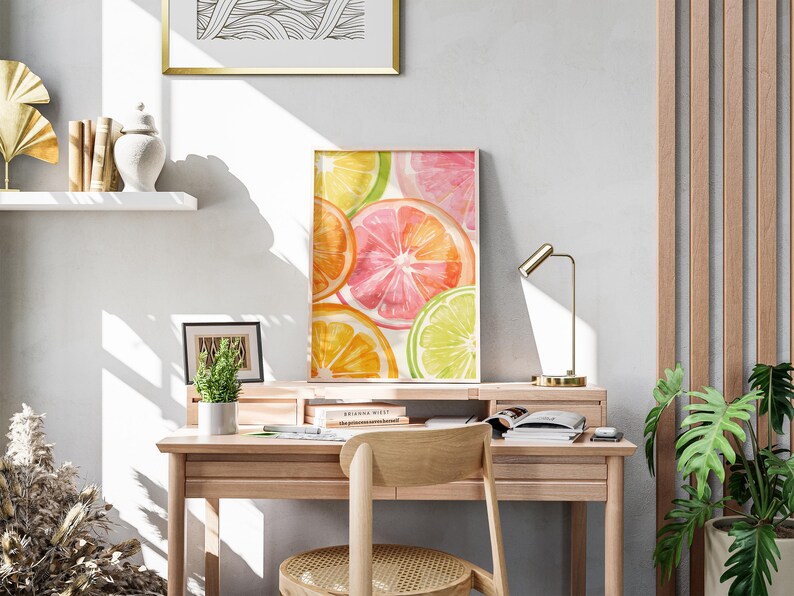 May include: A framed watercolor print of citrus fruit slices in shades of orange, pink, and green rests on a wooden desk. A small desk lamp and a potted plant are also on the desk. The room has a neutral color palette.