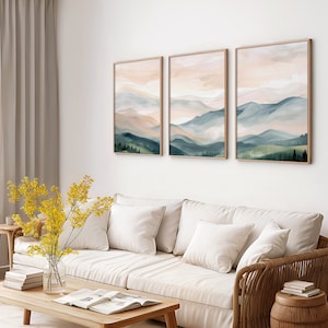 May include: Three abstract landscape paintings with a muted color palette of soft blues, greens, and pinks. The paintings are framed in a light wood frame and hung on a white wall above a white couch.