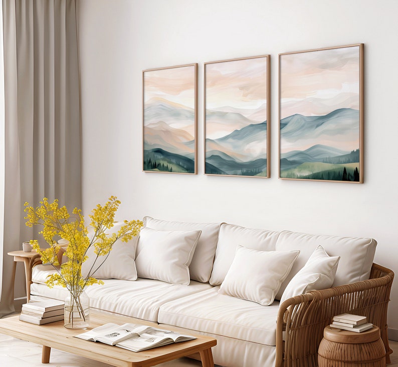 May include: Three abstract landscape paintings with a muted color palette of soft blues, greens, and pinks. The paintings are framed in a light wood frame and hung on a white wall above a white couch.