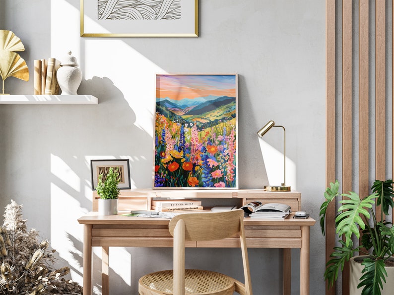 May include: A framed painting of a colorful wildflower meadow with a mountain range in the background. The painting is on a desk with a lamp, books, and a plant.