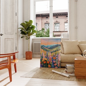 May include: A framed painting of a colorful wildflower meadow with a mountain range in the background. The painting is hanging on a white wall in a living room setting.