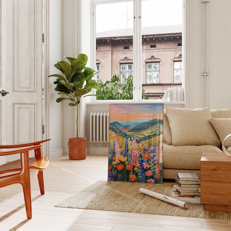 May include: A framed painting of a colorful wildflower meadow with a mountain range in the background. The painting is hanging on a white wall in a living room setting.