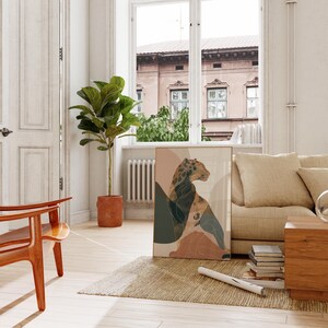 May include: A framed art print featuring an abstract leopard design in muted earth tones. The artwork includes shades of beige, brown, and green, with a minimalist aesthetic. The print is displayed in a living room setting with a sofa, rug, and wooden furniture.