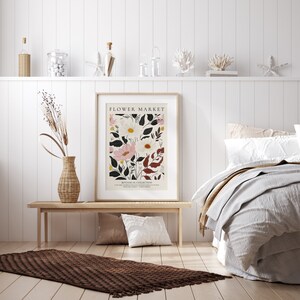 May include: A white framed poster with the text "FLOWER MARKET" and a botanical illustration of pink and white flowers with green and red leaves. The poster is hanging on a white wall above a wooden bench with a brown rug in front of it.