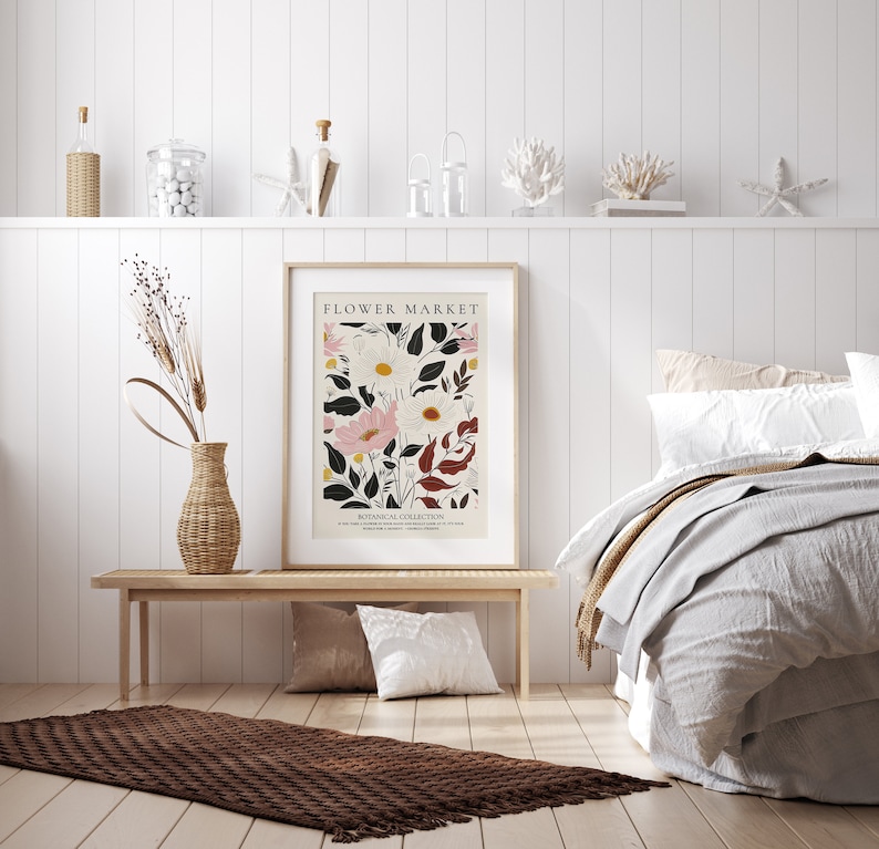 May include: A white framed poster with the text "FLOWER MARKET" and a botanical illustration of pink and white flowers with green and red leaves. The poster is hanging on a white wall above a wooden bench with a brown rug in front of it.