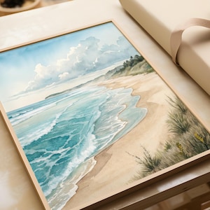 May include: A framed watercolour painting of a beach scene. The artwork shows a sandy beach, turquoise waves, and a blue sky with clouds. Green foliage is visible on the right. The frame is made of light-coloured wood.