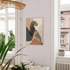 May include: A framed art print featuring a stylized leopard illustration. The artwork uses a palette of muted earth tones, including beige, brown, and green, with abstract shapes. The leopard is depicted in profile, with a spotted pattern. The print is displayed in a room with natural light.