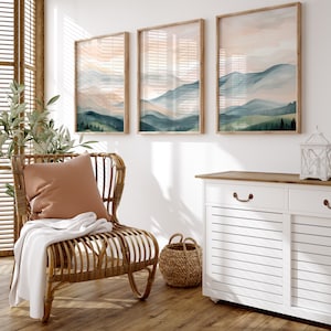 May include: A white cabinet with two drawers and two doors, a wicker basket, a rattan chair with a brown throw blanket and a pillow, and three framed watercolor paintings of mountains and a sunset.