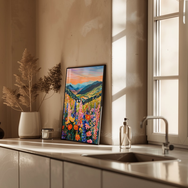 May include: A framed painting of a mountain landscape with a field of colorful wildflowers in the foreground. The sky is a vibrant orange and pink, and the mountains are a deep blue. The painting is framed in black.