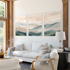 May include: Three framed watercolor paintings of a mountain landscape with soft pink and blue hues. The paintings are hung on a white wall in a living room setting with a white sofa, a white chair, and a black side table.