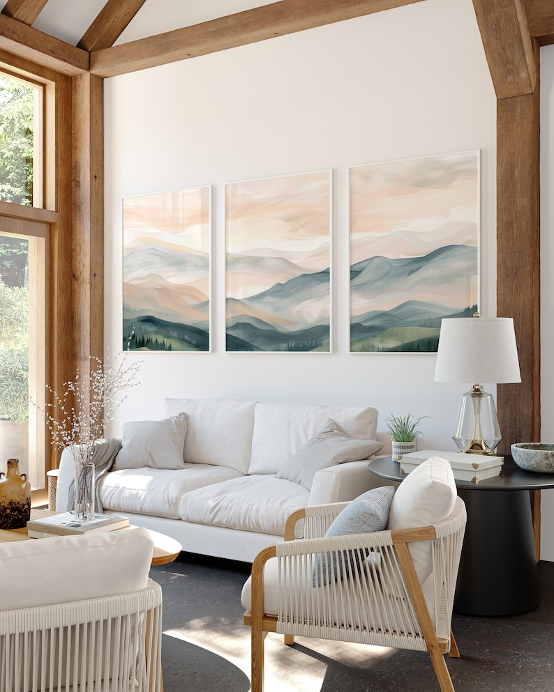 May include: Three framed watercolor paintings of a mountain landscape with soft pink and blue hues. The paintings are hung on a white wall in a living room setting with a white sofa, a white chair, and a black side table.