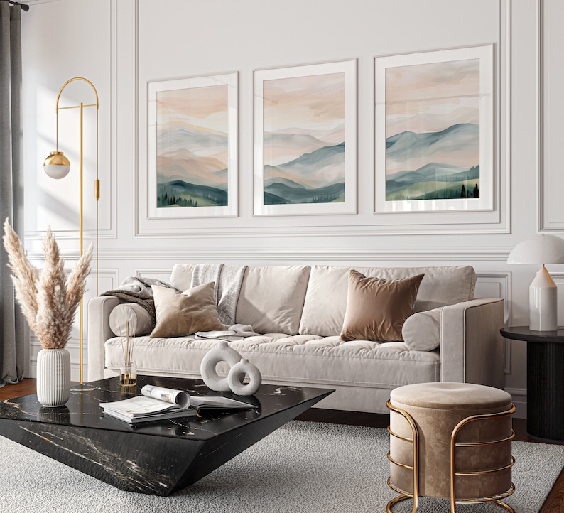 May include: A white sofa with three pillows, a black coffee table with a magazine on it, and a gold and beige ottoman. The sofa is in front of a white wall with three framed landscape paintings.